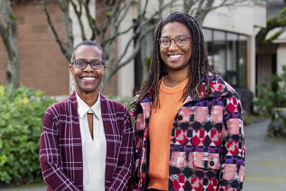 Two smiling Black women stand outside on the UVic campus grounds.