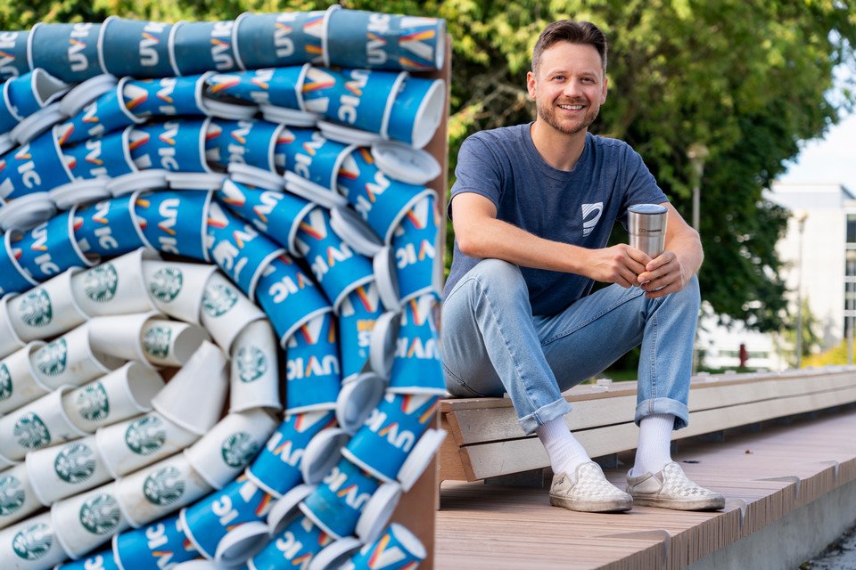 Matt Miller, UVic PhD student in Oceanography, behind a collage of UVic single-use beverage cups.