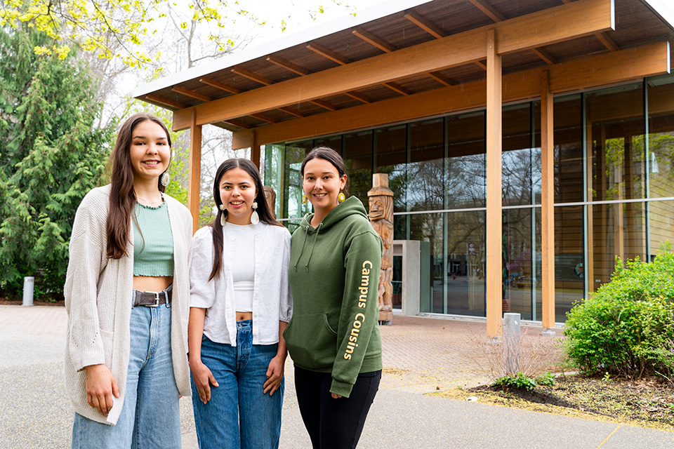 Three women stand in front of First Peoples House at the UVic campus.