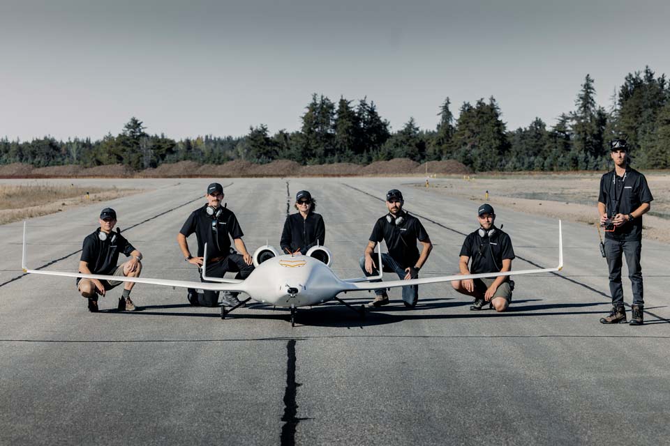 6 people sit or stand with a small white eco jet prototype on a runway.