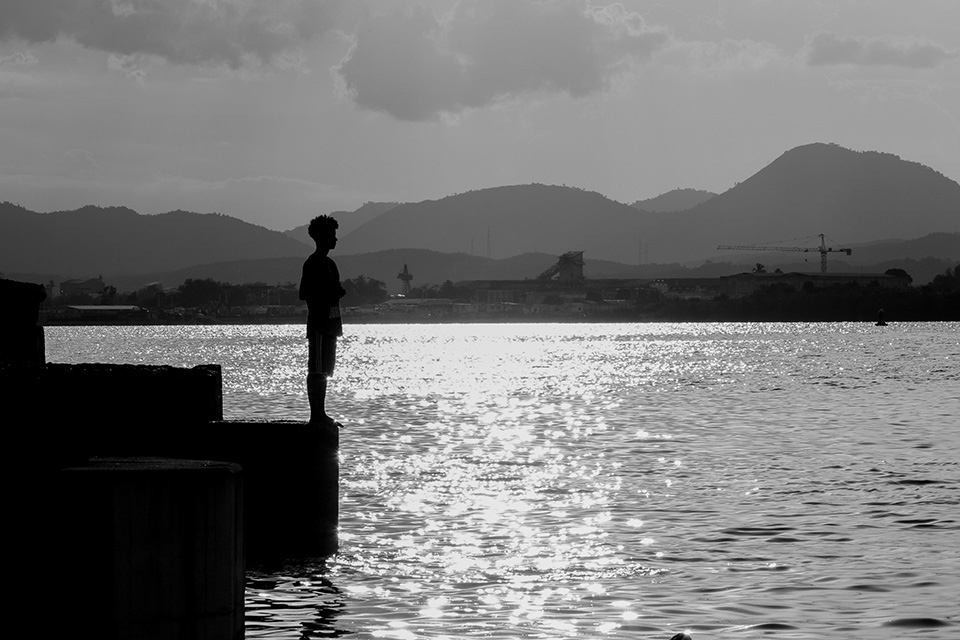 A black and white photo of a person standing at the edge of a platform at the water.