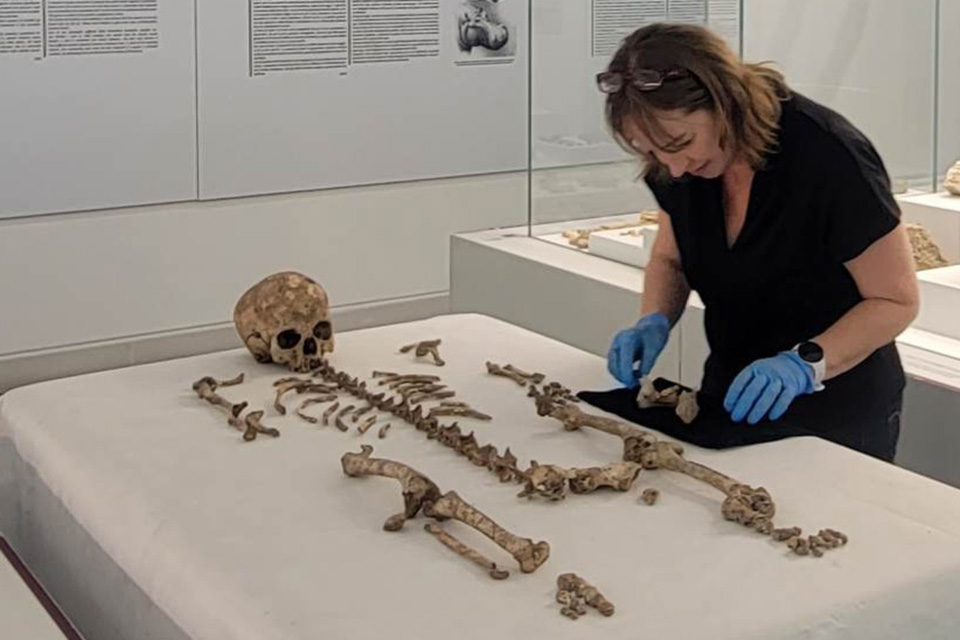 Female researcher wearing blue latex gloves bends over skeletal remains of a human laid out on a white table cloth. The remains include a skull, leg/arm bones, spine and other fragments.