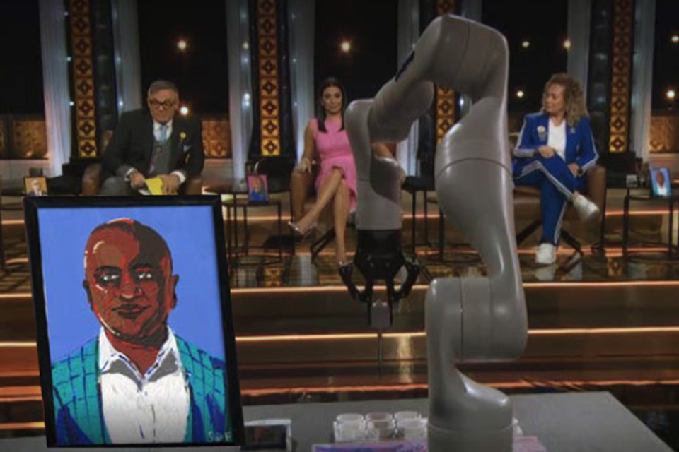 An illustration of a portrait of a man sits framed on a table beside a robotic arm. In the background, three people sit facing the table on the set of the tv show, Dragon's Den.