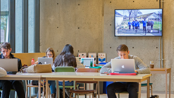 Students with laptops at desks in front of a digital screen.