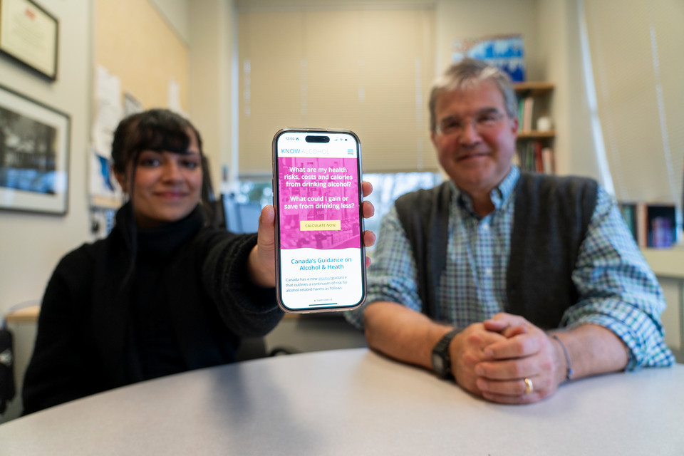 CISUR research associate Priya Johal and CISUR director Dr. Tim Naimi sit at a table. Priya holds a smartphone with the KnowAlcohol.ca website displayed.
