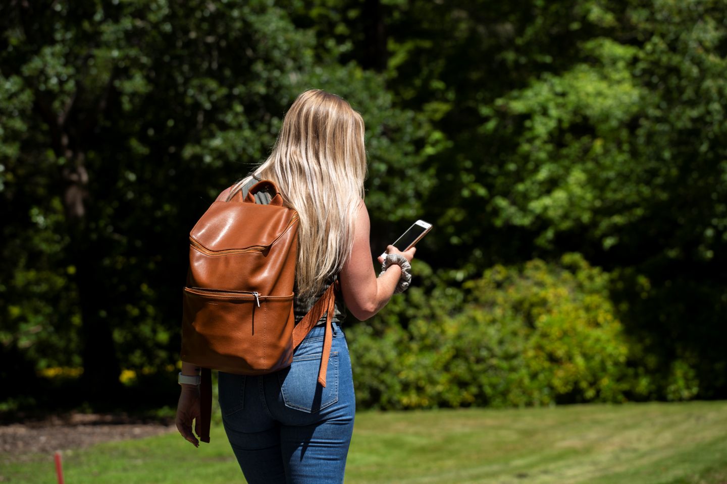 A student wearing a backpack and holding a smartphone walks across campus.
