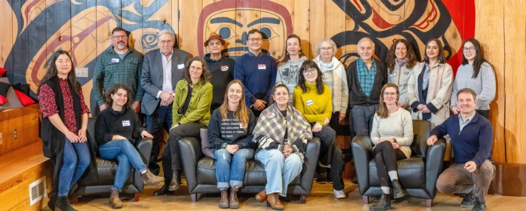 Eighteen individuals of various ages and backgrounds pose for a group photo beneath a mural painted in the traditional Haisla style.