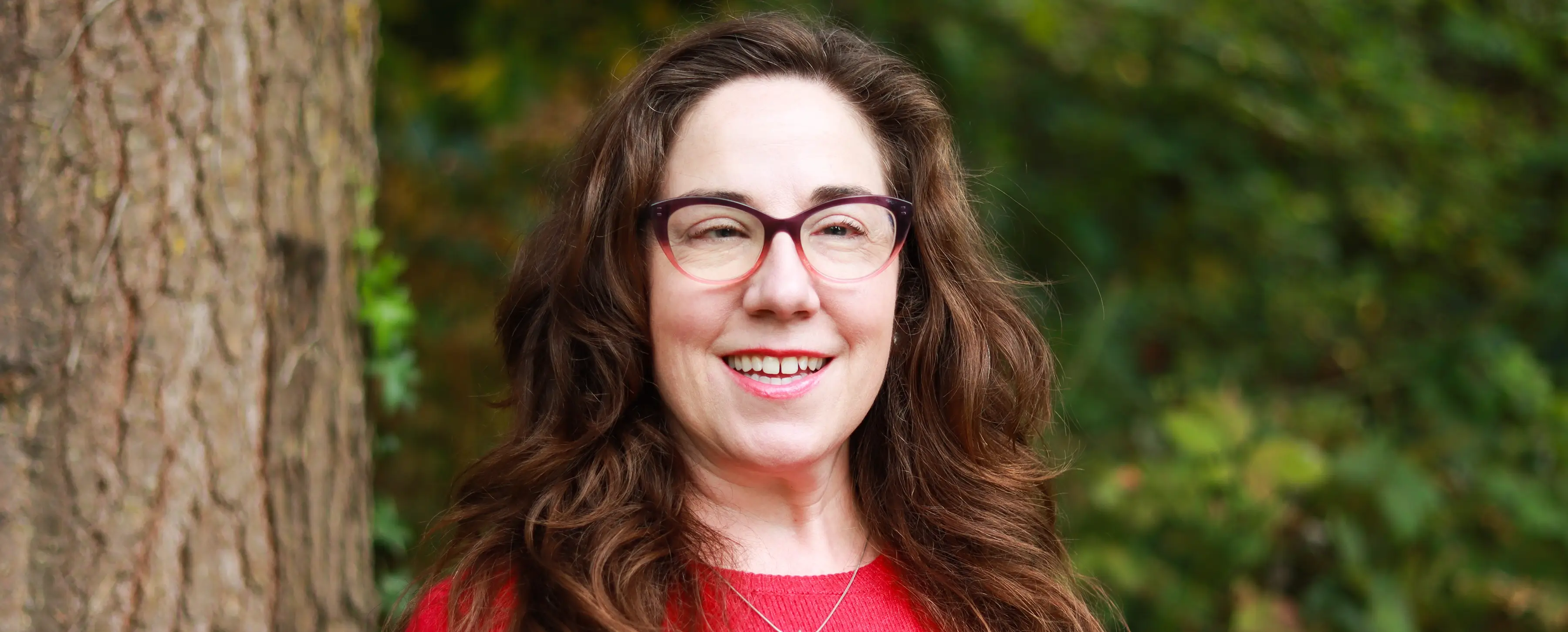 Headshot of Professor Rebecca Gagan, smiling and wearing a red shirt with blurred forest in background.