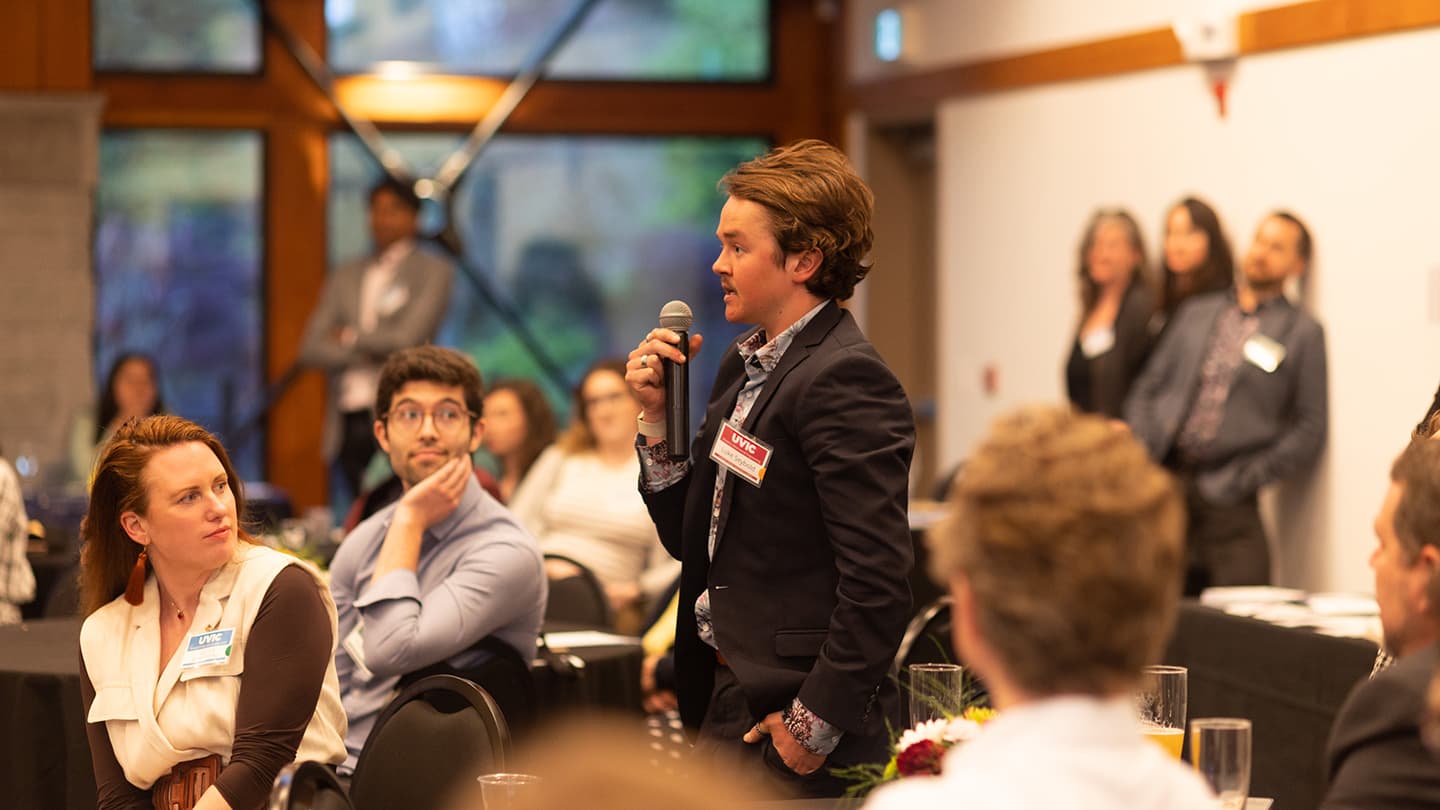 A standing man holds a microphone, asking a question, in a room full of people.