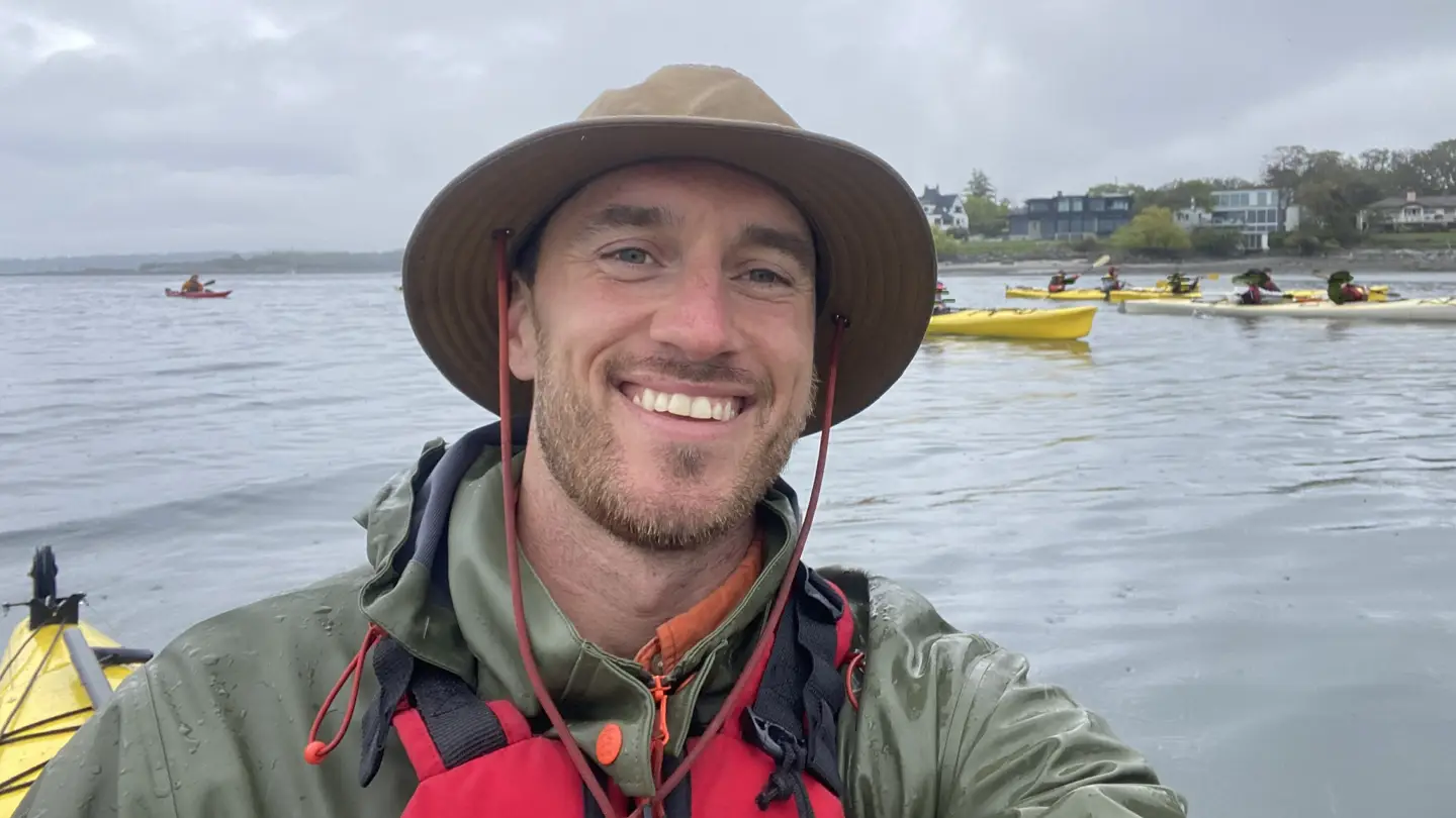 A student smiling in a kayak with kayaks and ocean in the background.