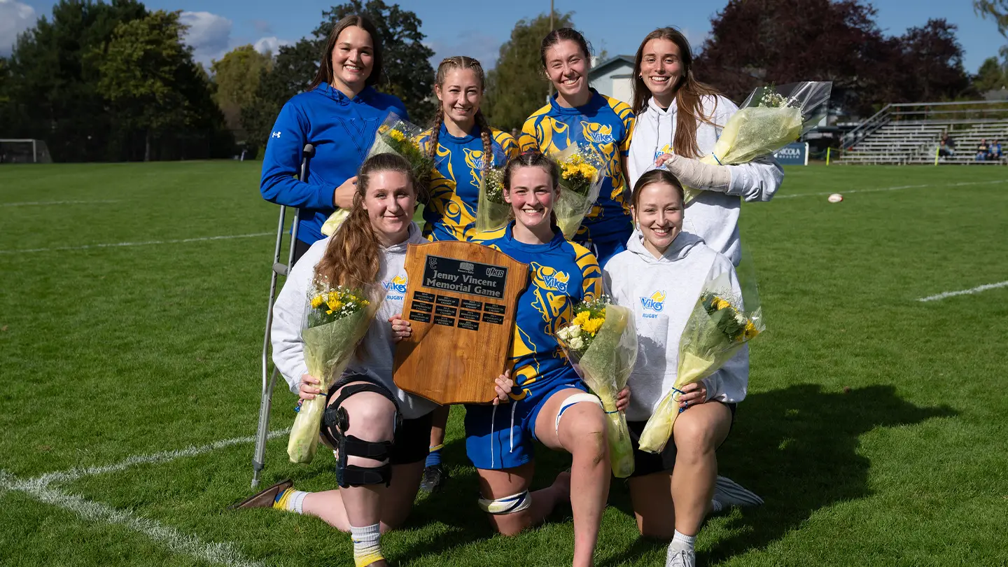 A smiling group of teammates hold a plaque in a sunny rubgy field.