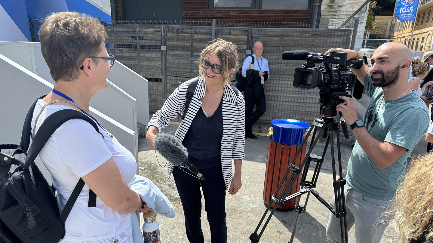 A woman is interviewed by two journalists: one is holding a microphone and another is behind a video camera.
