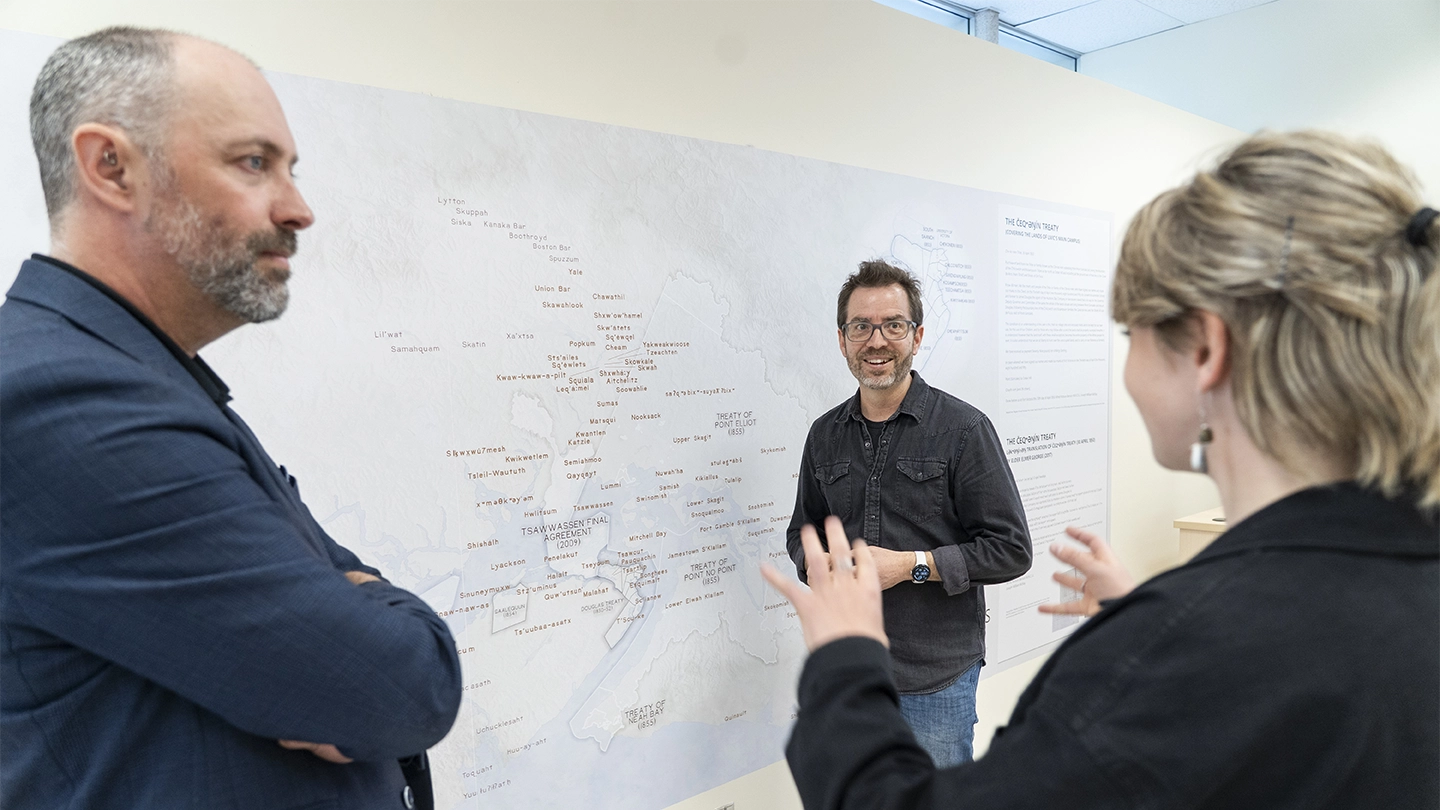 Someone gestures with their hands as two people listen to them, with a map on the wall in the background.
