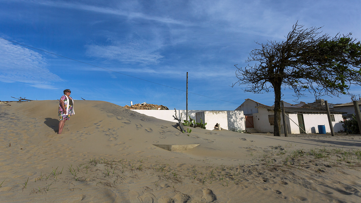 A woman stands on a hill of sand that is engulfing her home in the background.