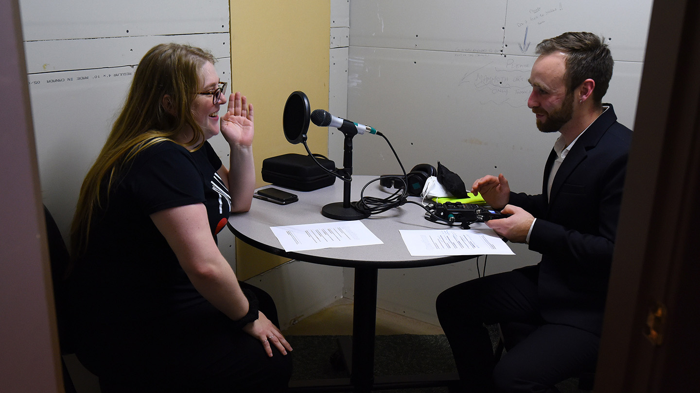 A woman sits in front of a microphone interviewing a man with notes in front of them.