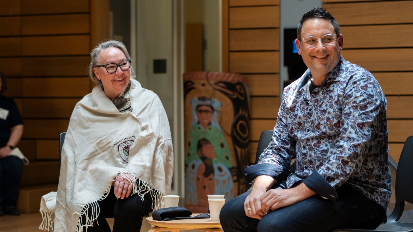Siila Watt-Cloutier and Dr. Ian Mauro sit side by side in chairs at the ceremonial hall in First Peoples House at UVic. Both are smiling and looking to the right, engaged with someone in the audience. Watt-Cloutier is wrapped in a ceremonial blanket, and Mauro wears a floral shirt. Both wear glasses. A small table with three cups of water sits between them.