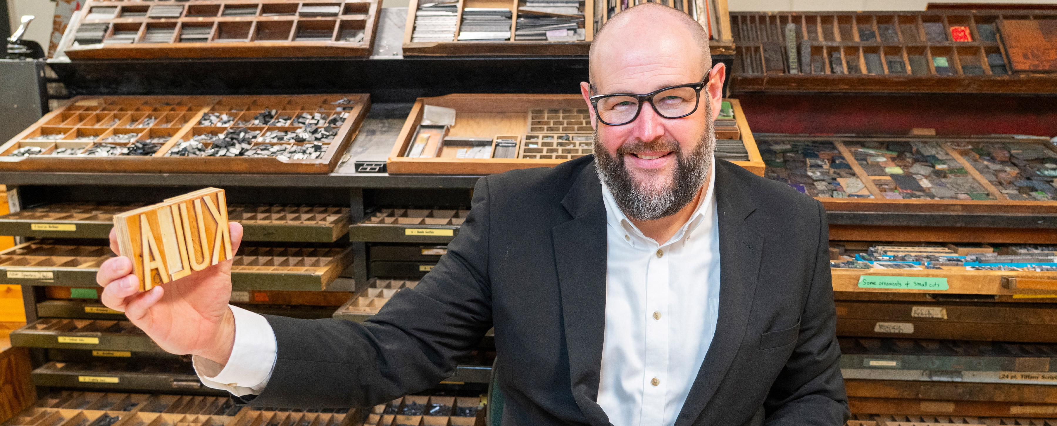 Matt is holding the KULA word in block letterpress letters in his right hand, and placing his left hand on a wooden tray of metal type; behind him are numerous trays of typefaces in wood blocks and metal.