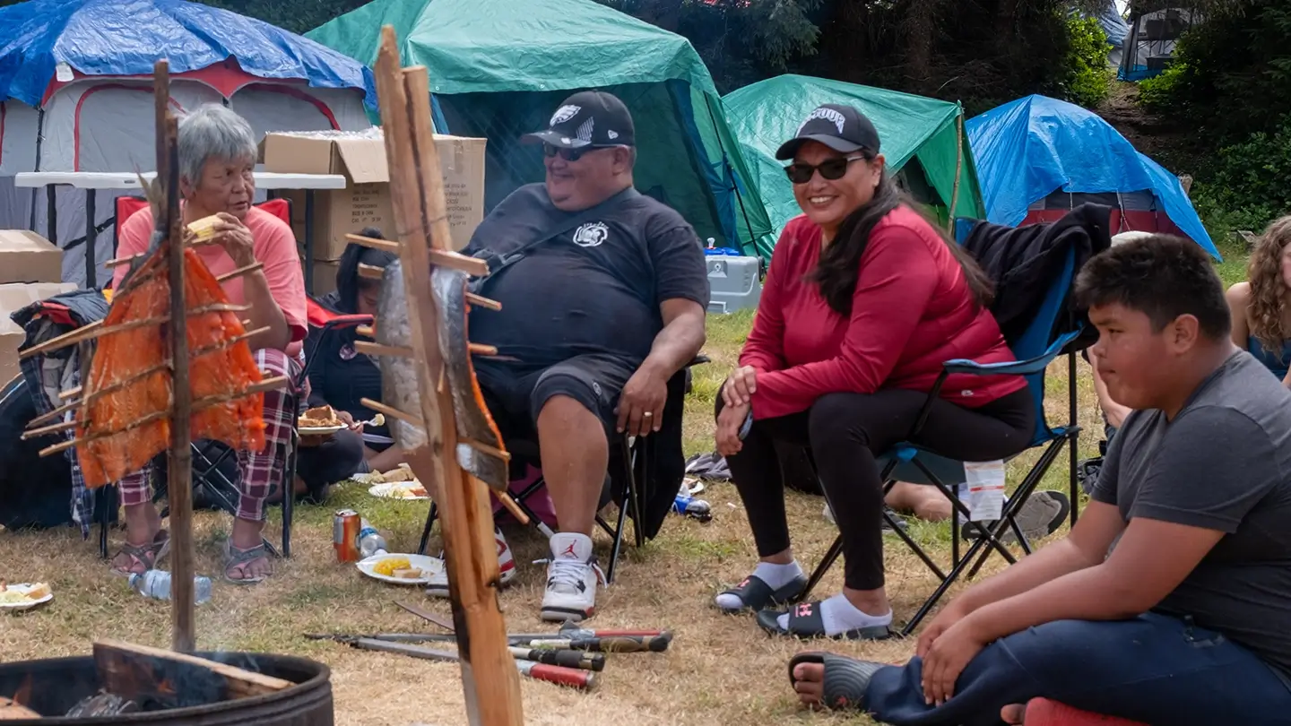 A small group of people sit and relax around an outdoor fire where fish fillets are being cooked on wooden racks. Tents are visible in the background. A gray-haired woman eats while others smile and socialize, including a man in a black shirt and cap, a woman in a red jacket and sunglasses, and a boy sitting cross-legged on the grass.