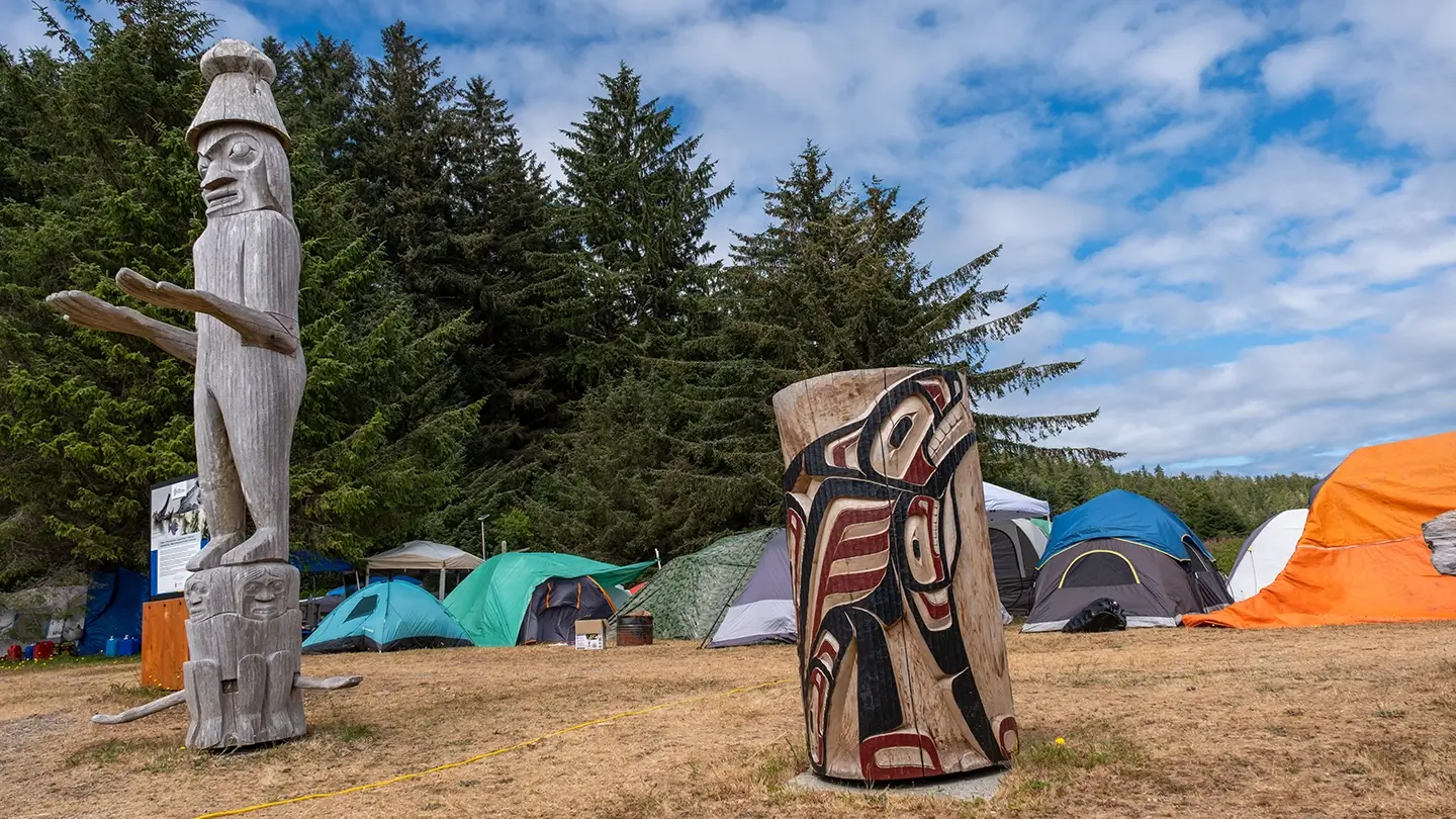 A large wooden Mowachaht-Muchalaht carving of a human figure with outstretched arms stands on a grassy area near a forest. In the foreground is another carved log featuring a painted design of a stylized animal in red, black, and white. Behind the carvings, multiple colorful tents and tarps are set up in a campsite under a partly cloudy sky.