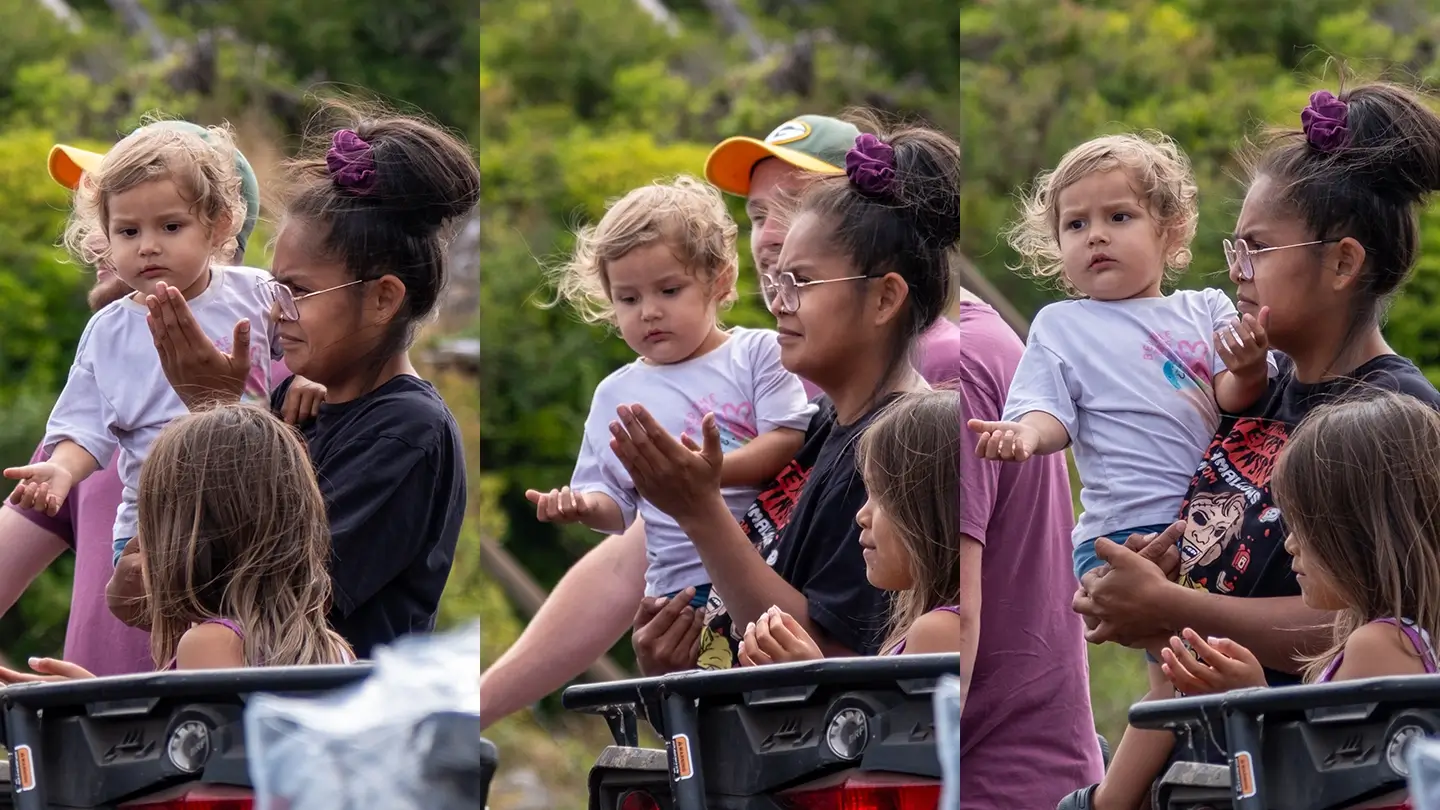 A series of three side-by-side photos shows a young Indigenous woman holding a small child at an outdoor event. The woman raises her hands in a gesture of welcoming, while the child looks on curiously before raising her own hands. Another child stands nearby, also watching. Trees and a crowd are visible in the background.