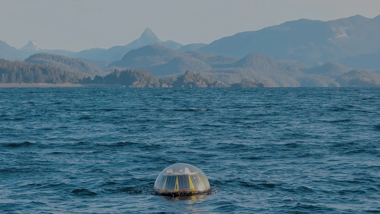 A wave resource assessment buoy with a clear dome top and yellow base floats on deep blue ocean water. In the distance, forested islands and layered mountain ranges rise under a hazy sky, creating a rugged coastal backdrop.