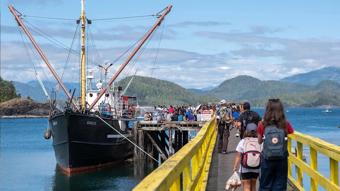 A crowd of people walk along a yellow-painted wooden dock toward a large black ship docked at the end, with forested hills and blue water in the background. Some carry backpacks or bags, and the dock is busy with activity under a bright, partly cloudy sky.