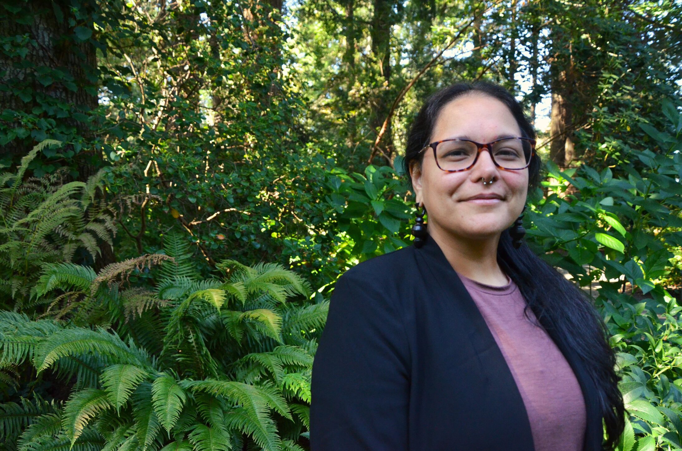 A woman with glasses and long dark hair stands outdoors, smiling softly. They're wearing a purple top and black blazer. Lush green foliage forms the backdrop. 