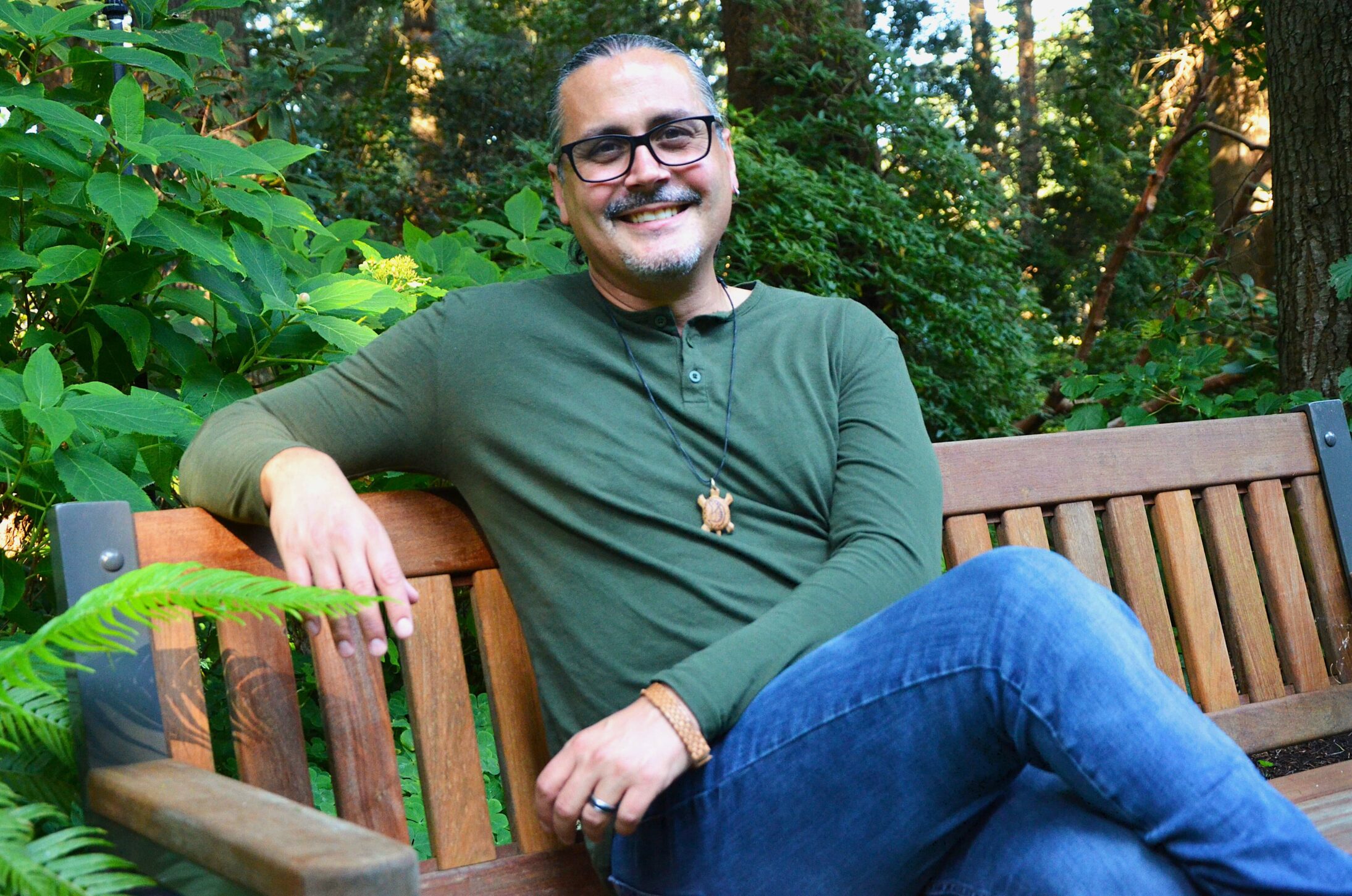 A smiling man with glasses and a mustache sits on a wooden bench in a lush green garden. They wear a green shirt and jeans, exuding a relaxed, happy vibe. 