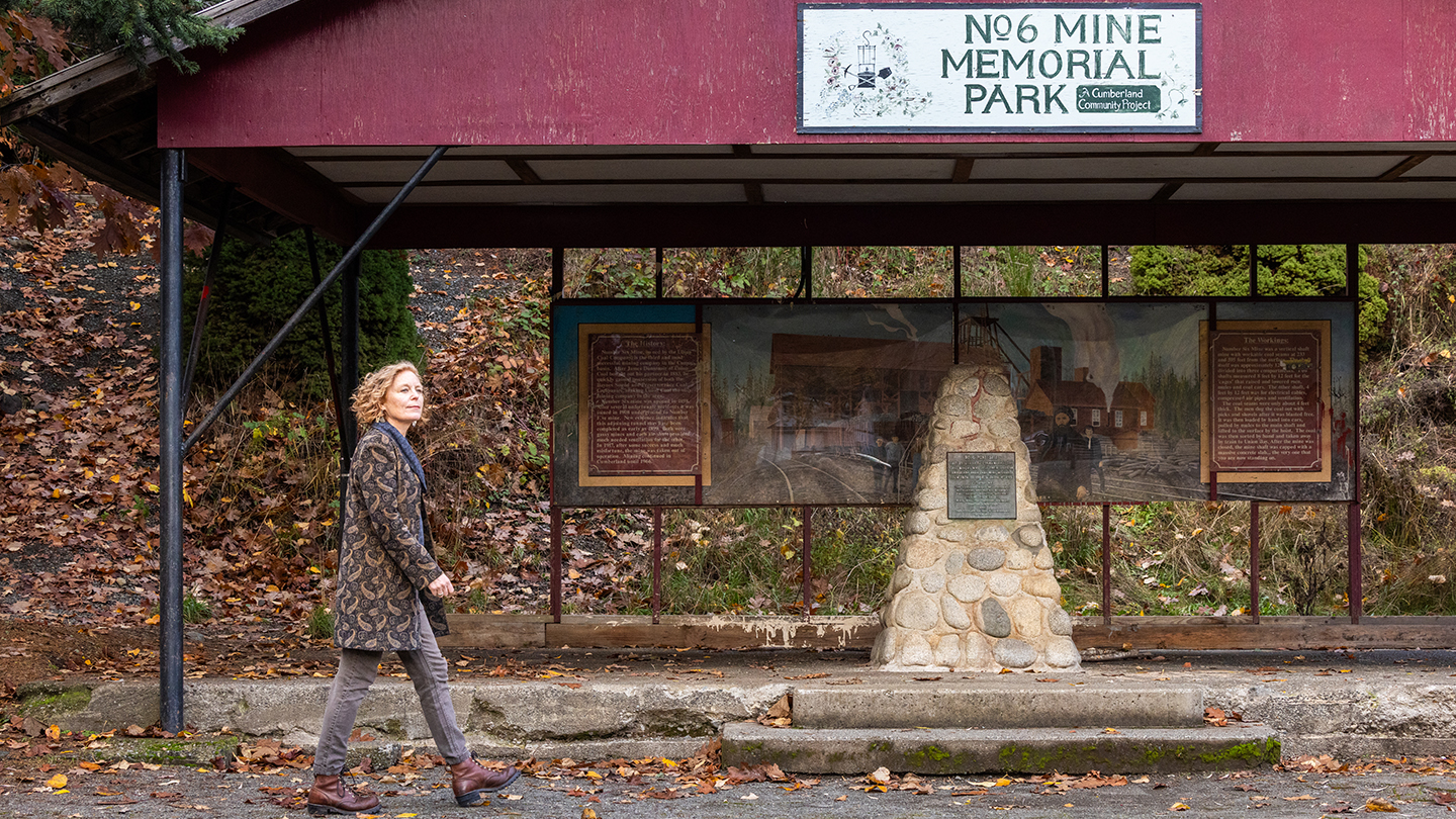 A person walks past a covered outdoor memorial display at a former mine site. The structure includes a stone monument with a plaque and illustrated panels showing historical scenes. Fallen autumn leaves cover the ground and trees line the background.