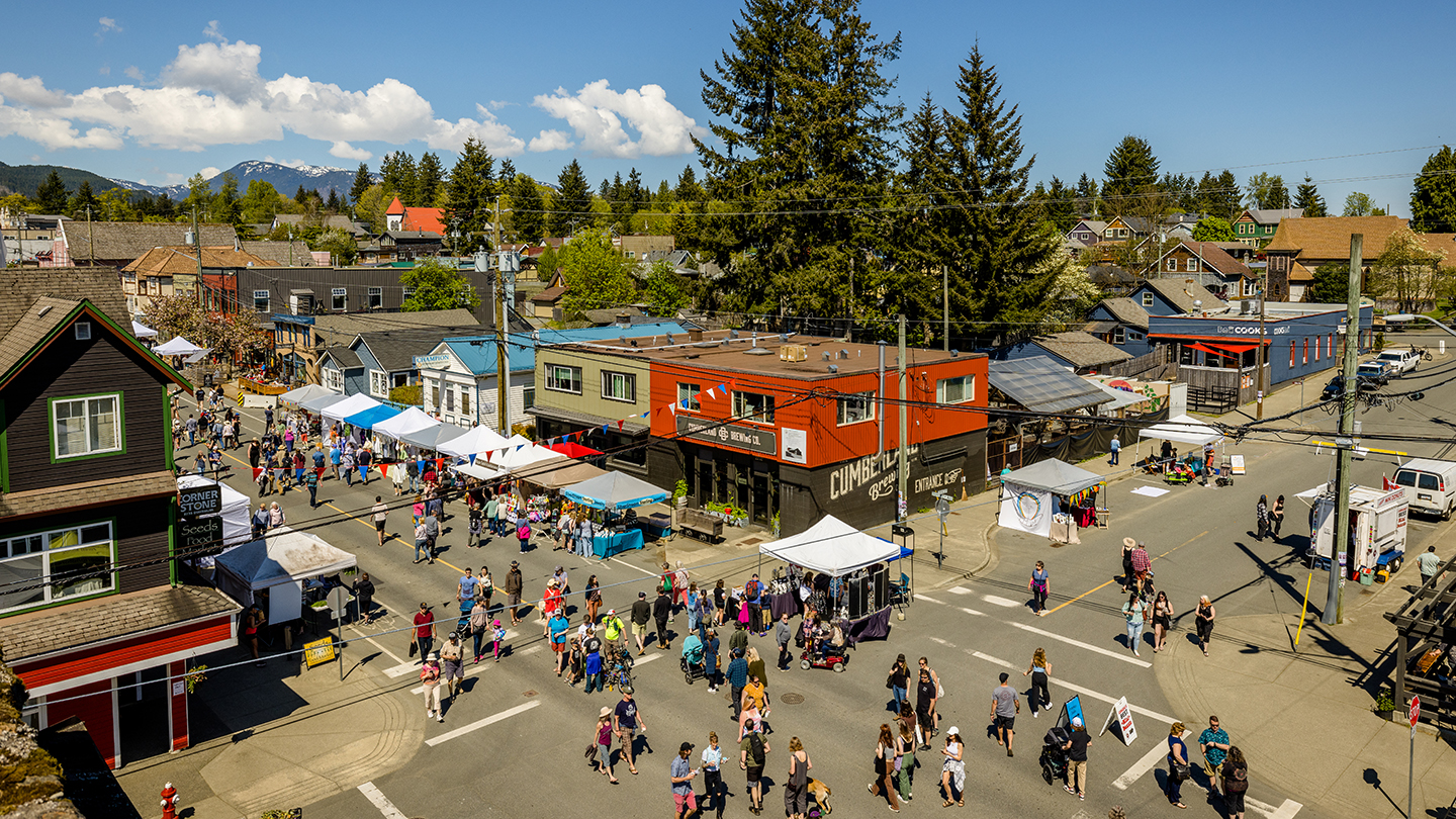 An aerial view of a lively street festival in Cumberland. Crowds walk among market tents and vendor booths set up along intersecting streets. Colourful buildings, tall trees, and distant mountains appear in the background under a clear blue sky.