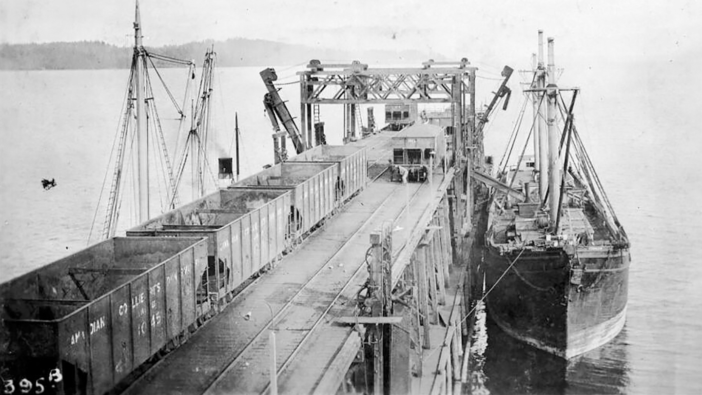An old black and white photograph of rail carts filled with coal being at Union Bay Wharf in Cumberland. The coal awaits loading onto an old industrial steam ship that is docked alongside the wharf.