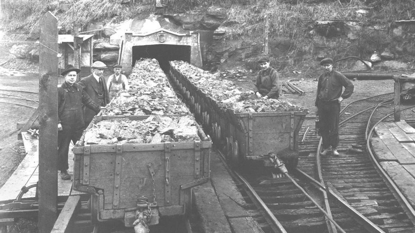 An old black and white photograph of five men standing alongside coal-filled rail carts coming out of a mine shaft behind them.