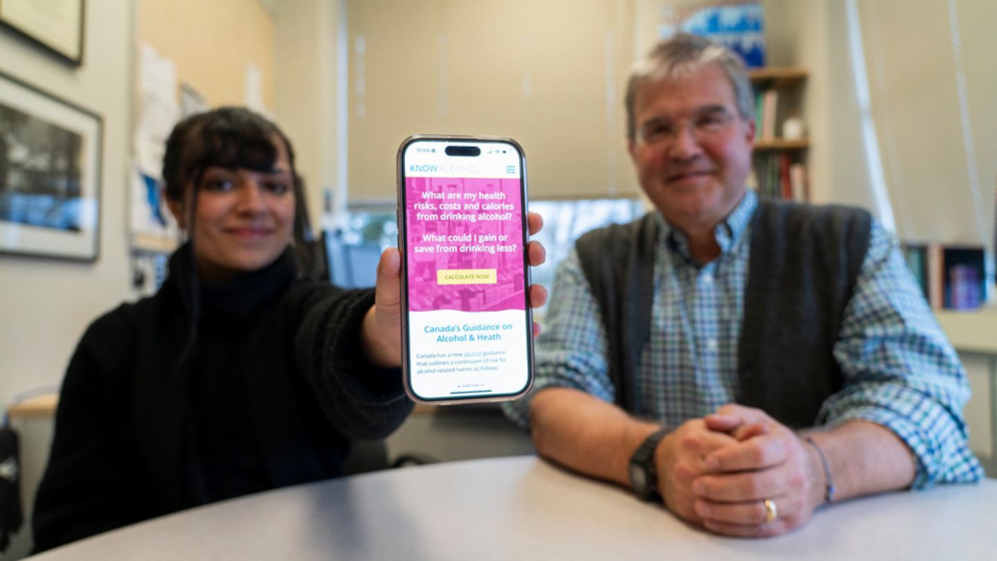 CISUR research associate Priya Johal and CISUR director Dr. Tim Naimi sit at a table. Priya holds a smartphone with the KnowAlcohol.ca website displayed.