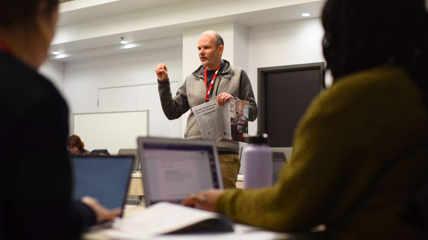 A man stands and speaks to a group in a classroom while holding papers; two people in the foreground are seated with laptops and notebooks.