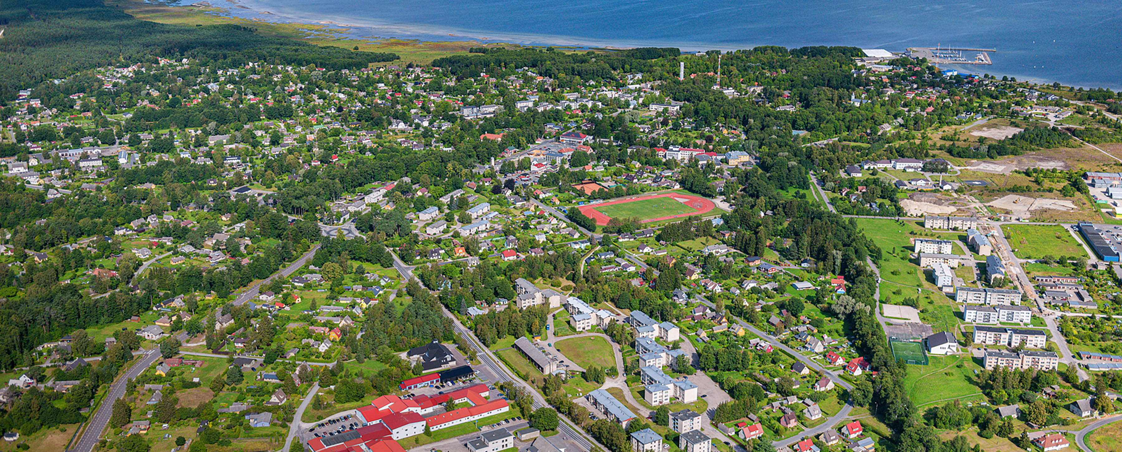 Aerial view of Kärdla, Hiiumaa County, Estonia, showing a small coastal city surrounded by forest, with residential neighbourhoods, community buildings, and the Baltic Sea coastline in the background.