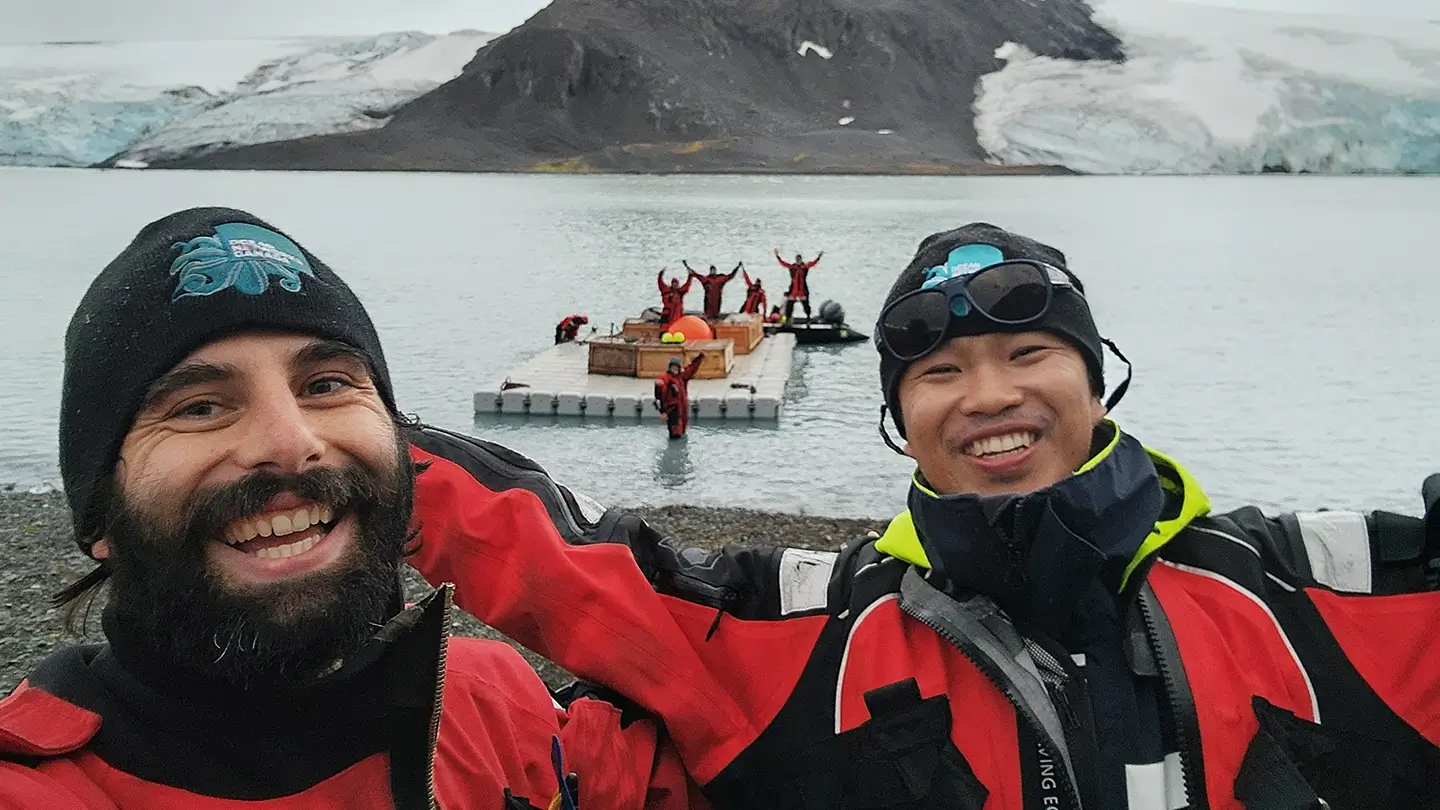 Two men wearing toques and red jackets pose together on a rocky shore. Behind them a group of people stand in the water and on a dock.