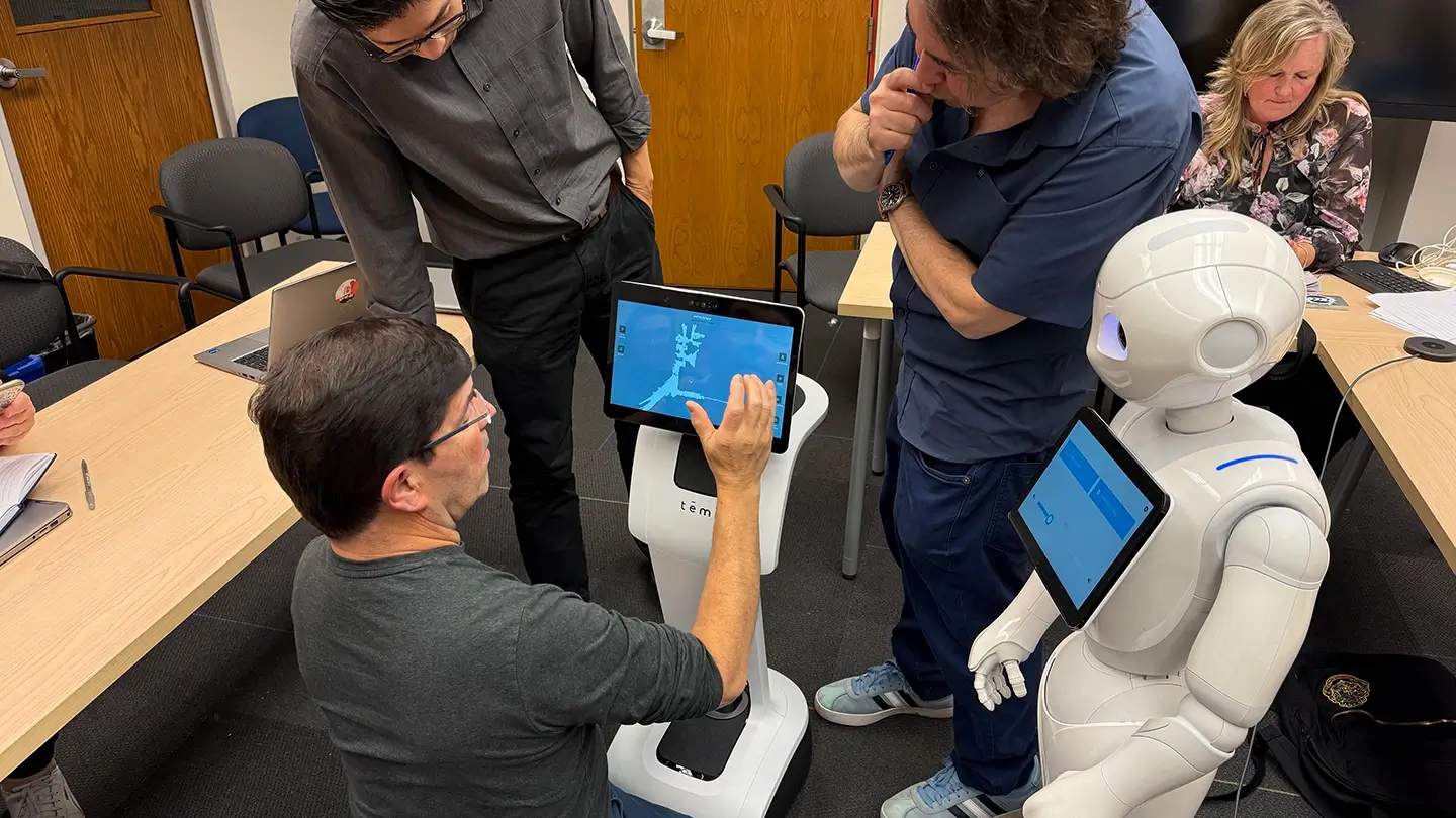 A Pepper robot on the right. A man's hand is programming a second robot on the left.
