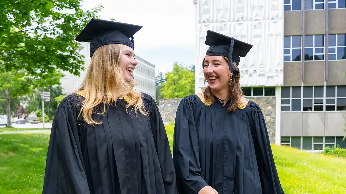 Camryn Thompson and Maya deWitt wear graduation caps and gowns and smile at each other.