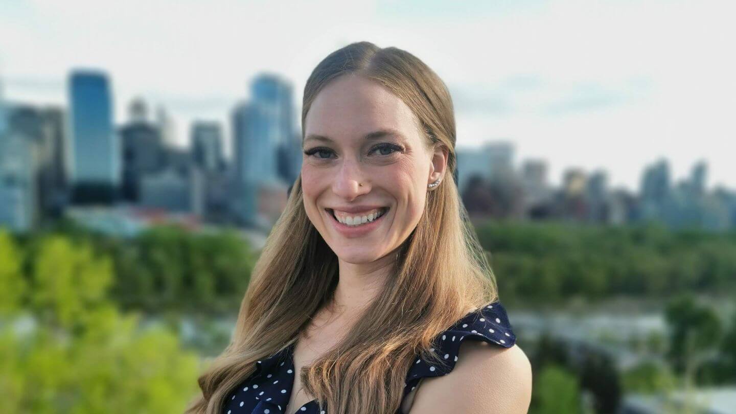 A young woman with blonde hair stands smiling in front of a city landscape