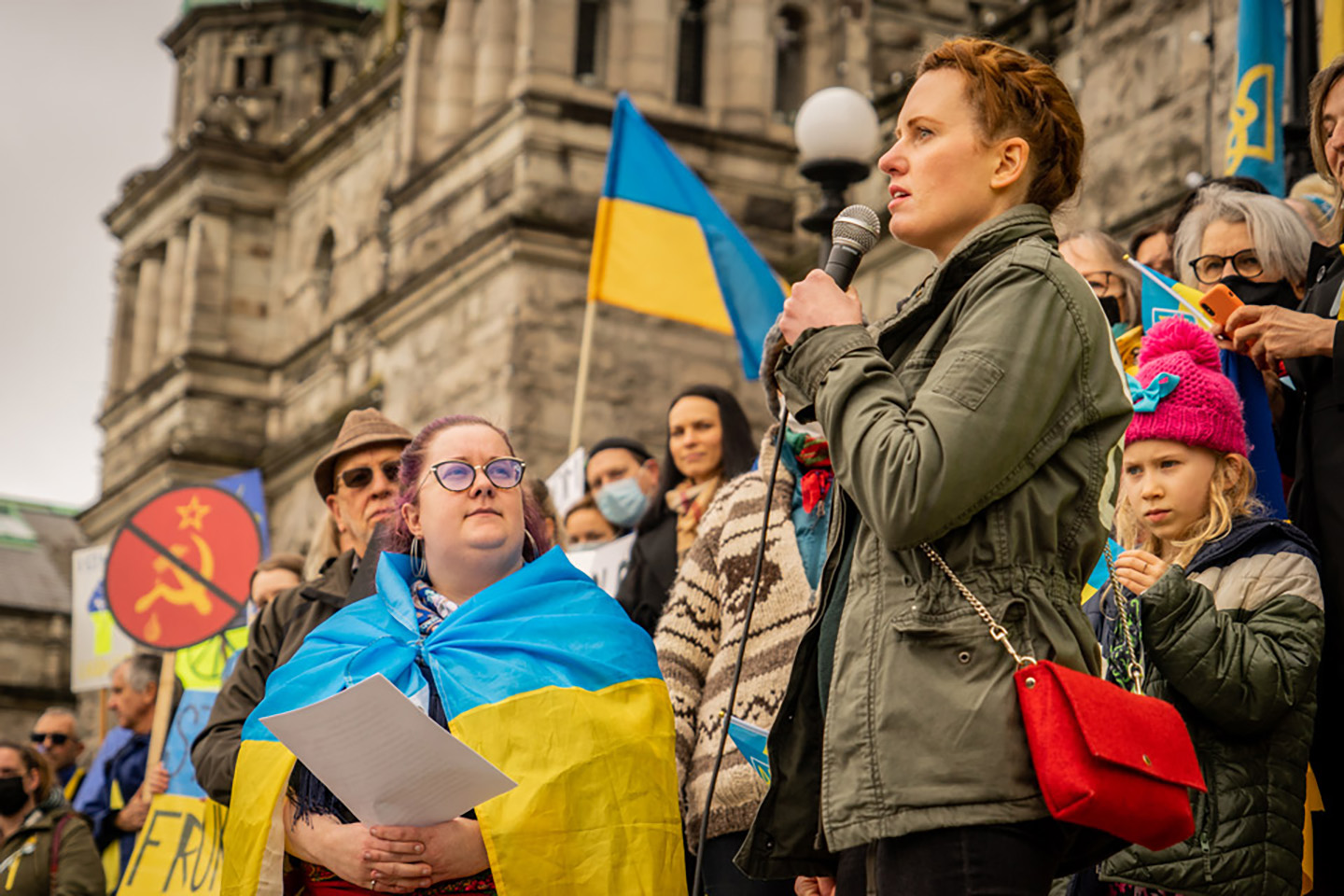 Woman with red hair talks into a microphone in front of a crowd of people carrying and wearing Ukrainian flags, assembled in front of Victoria's Legislative Assembly building.
