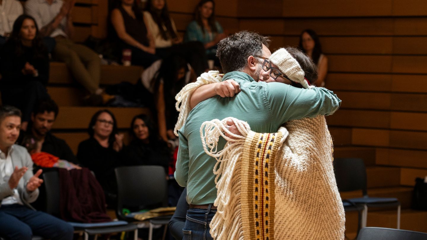 Indigenous woman wrapped in a woven blanket embraces another person during a ceremony.