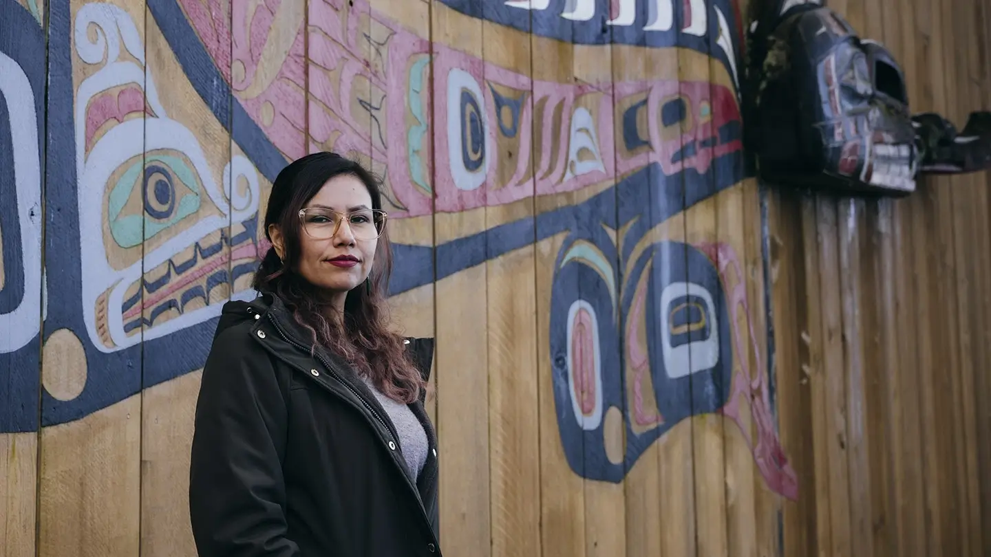 A woman wearing glasses and a dark coat stands beside a wooden wall featuring colourful Indigenous Northwest Coast-style artwork. Carved masks hang in the background as she looks toward the camera.