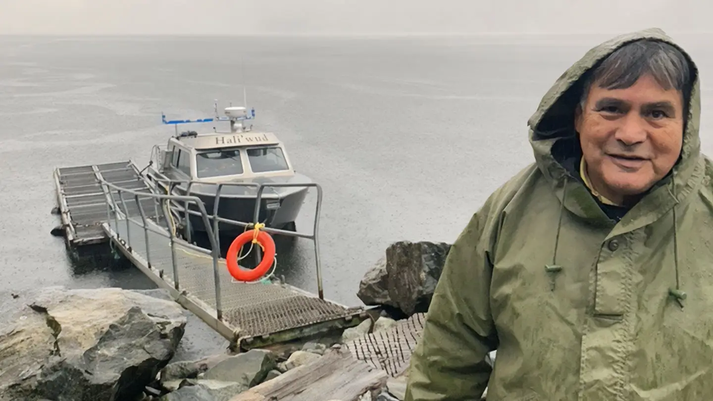 A man in a green rain jacket stands beside a small dock and boat on a rainy shoreline, with calm grey water and rocky terrain in the background.