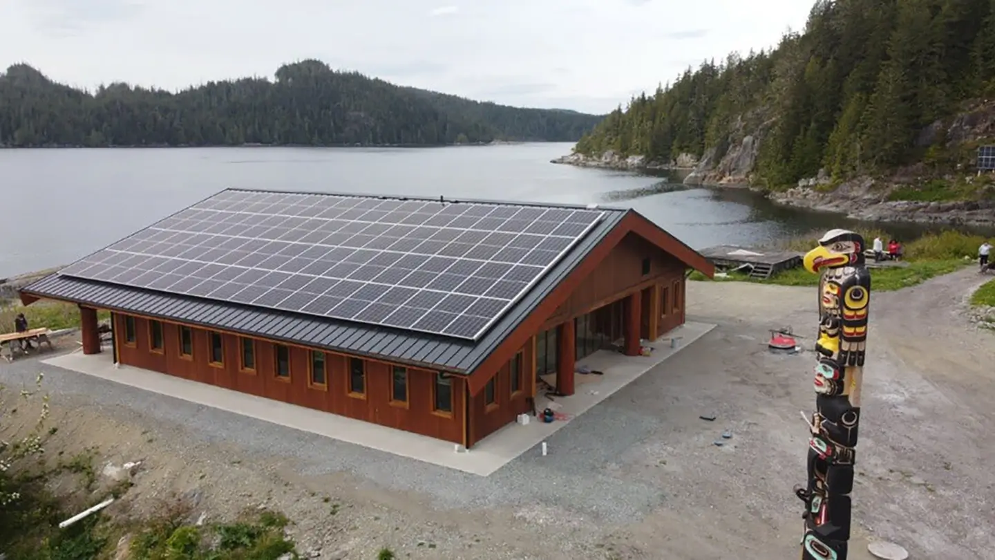 A large wood-clad building with a roof covered in solar panels sits beside a calm inlet surrounded by forested hills. A tall carved totem pole stands near the shoreline and gravel road in the foreground.