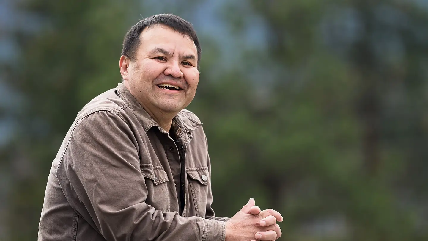 A smiling man wearing a brown jacket stands outdoors with his hands clasped, with blurred green forest scenery in the background.