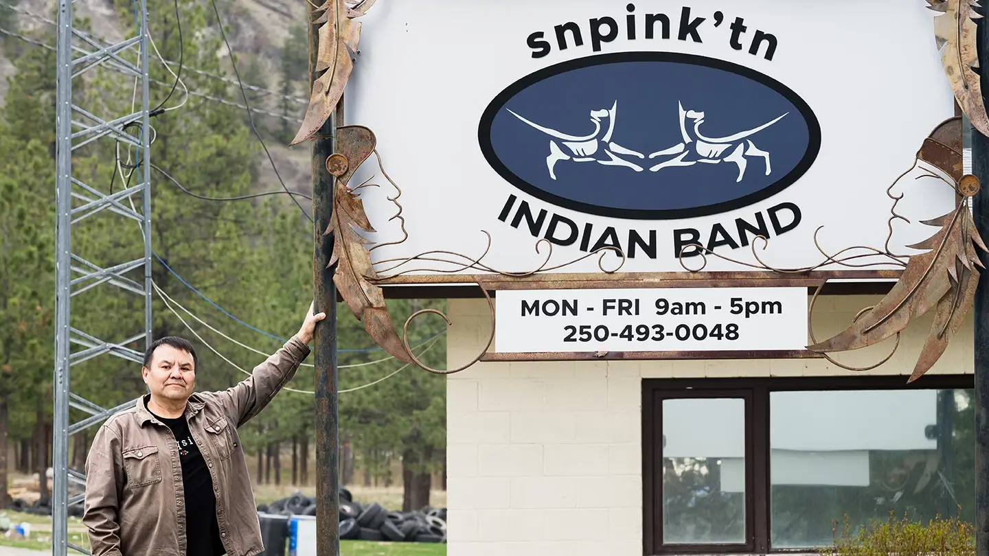 A man stands beside a white and blue sign reading “Snpink’tn Indian Band” with office hours and phone number displayed below, outdoors near trees and utility poles.