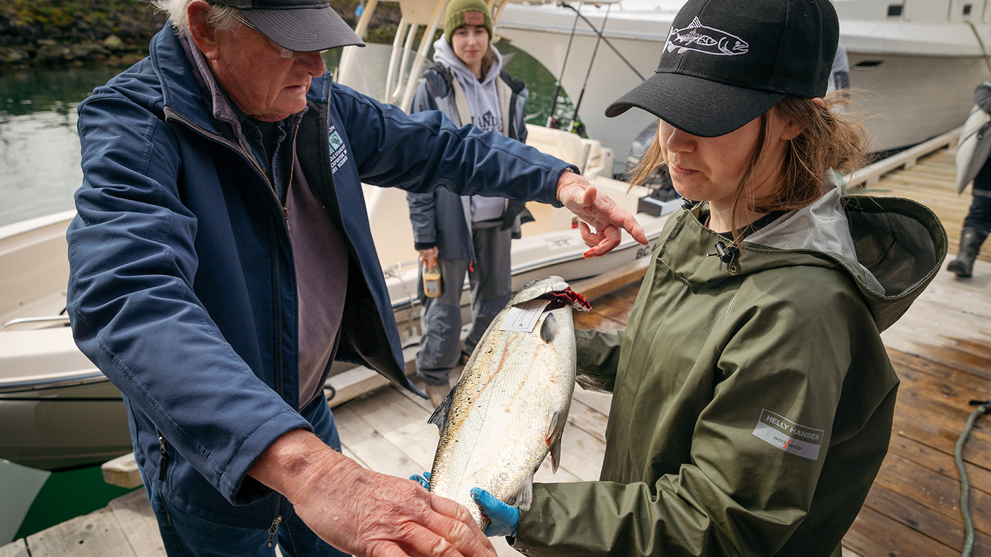 Fisherman with grey hair teaches a young person wearing a ball cap about fish as they hold a Chinook salmon in their arms.