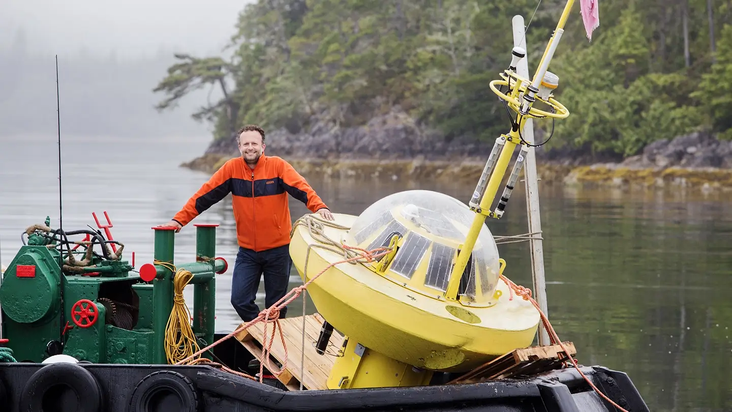 Brad Buckham stands with scientific equipment on a vessel on the water. Trees along a coast line are in the background.