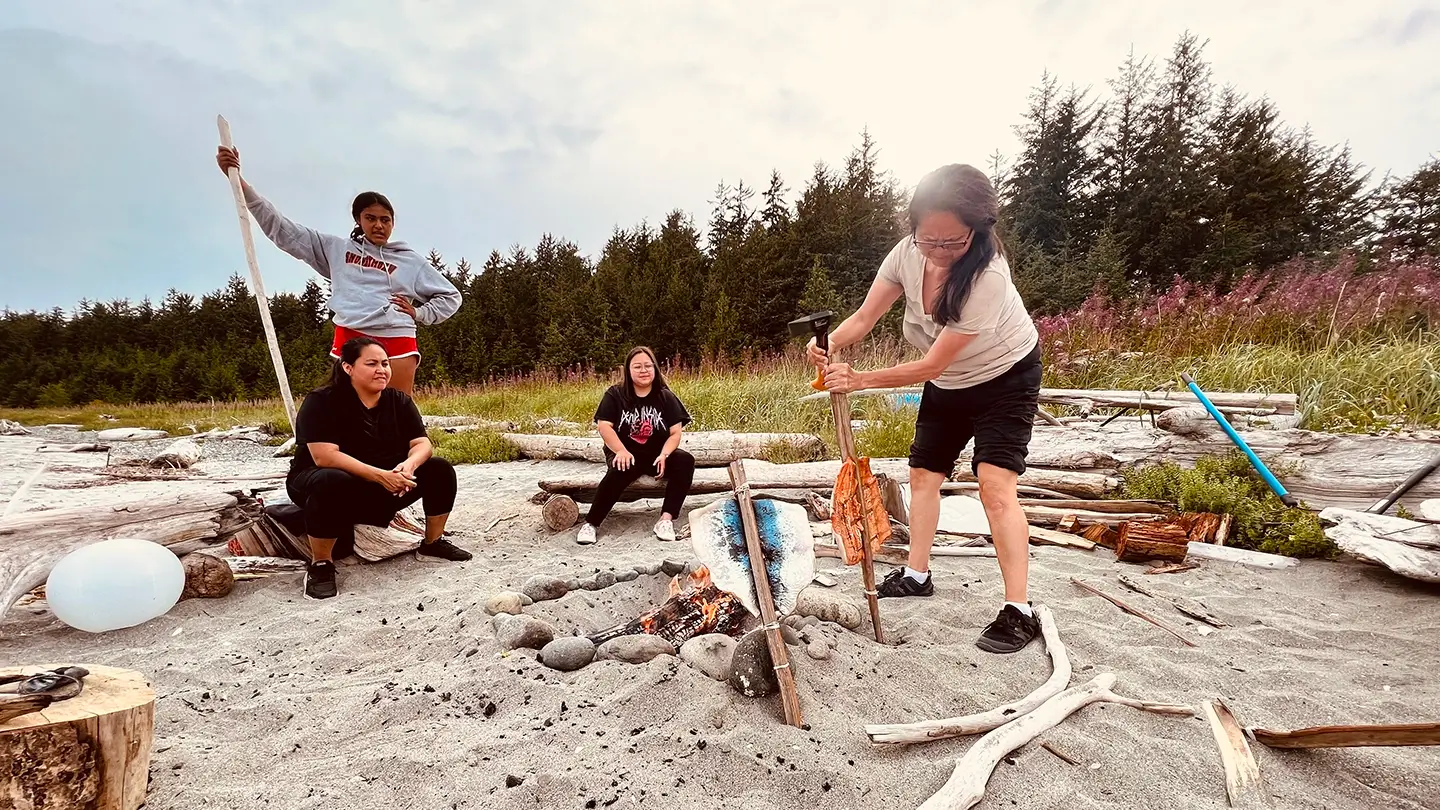 Four people sit and stand around a small fire as one person cooks salmon.