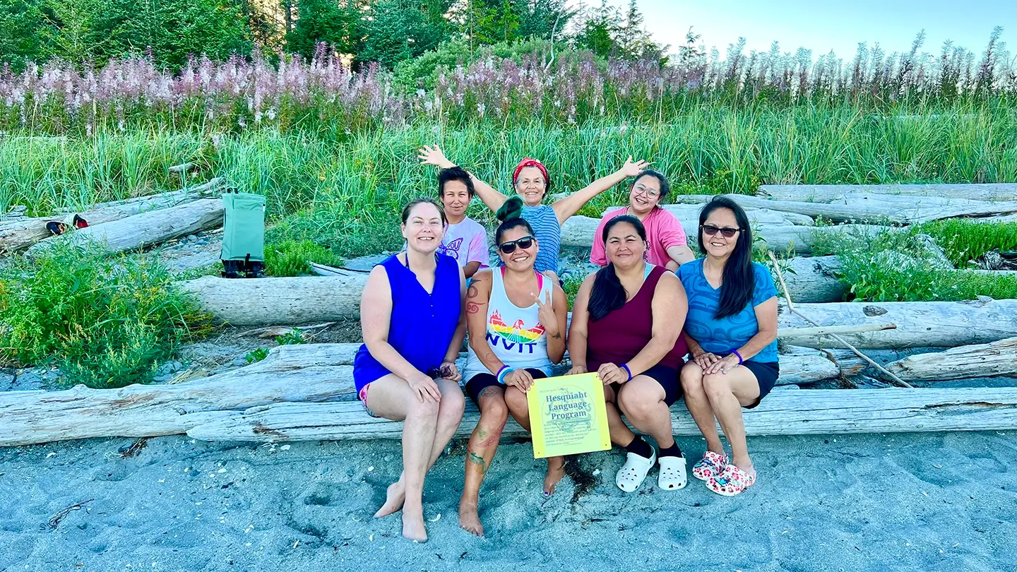 Seven people sit on a log at the beach with fireweed growing in the field behind them.